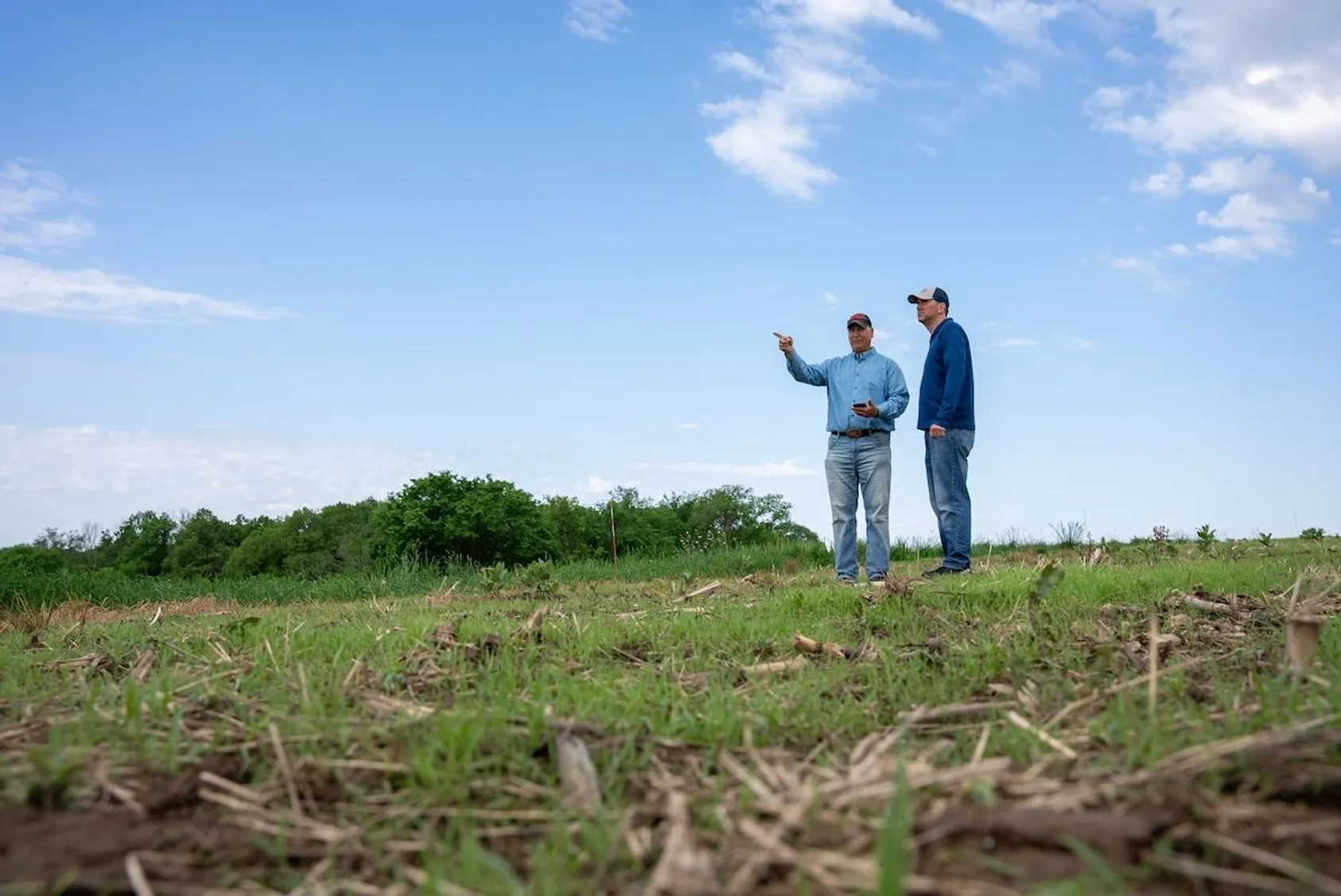 Two men standing in open field or pasture area under blue sky, appearing to discuss or evaluate the rural property