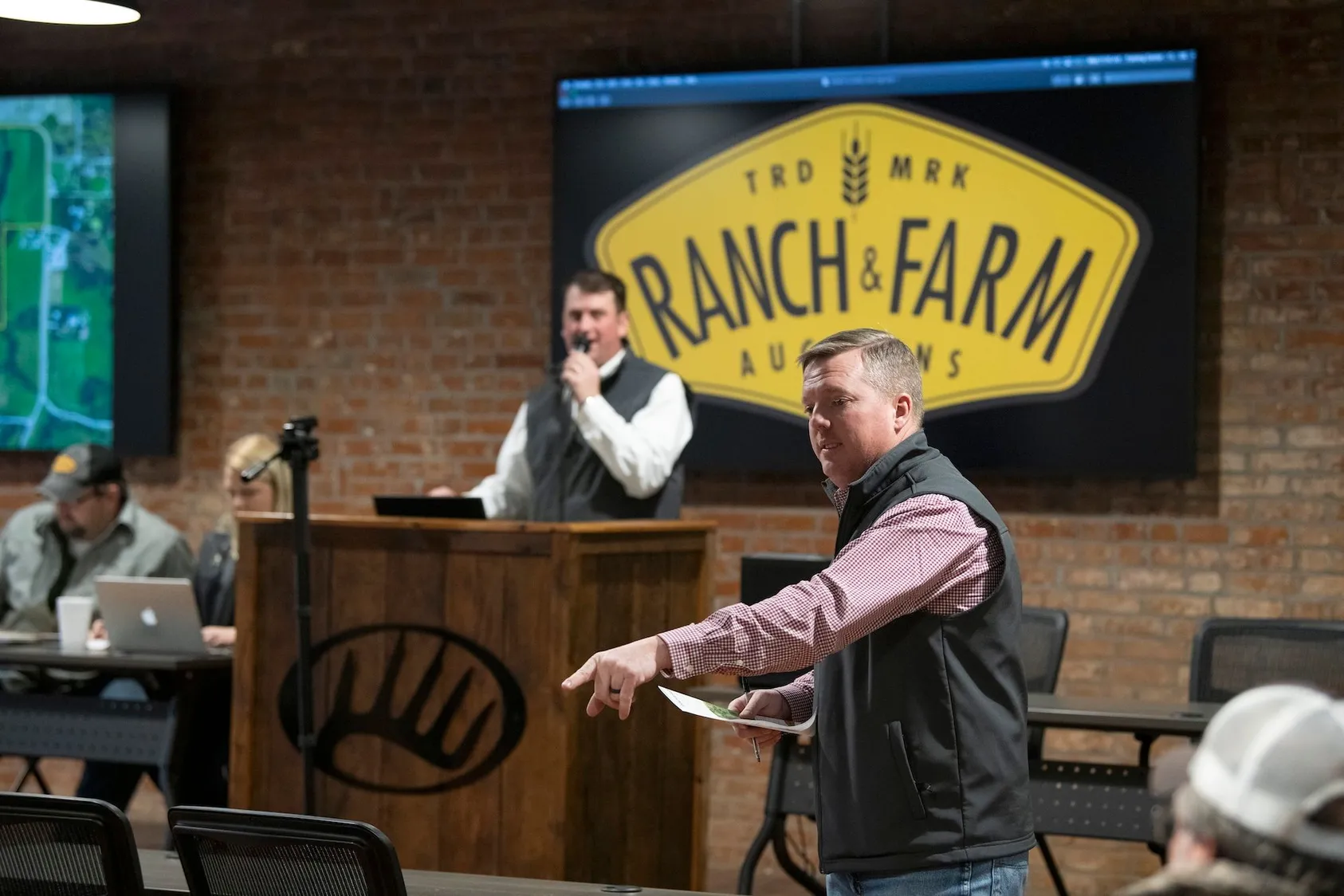Auctioneer at podium conducting land auction under 'Ranch & Farm Auctions' sign in brick venue, showing property sales and investment opportunities
