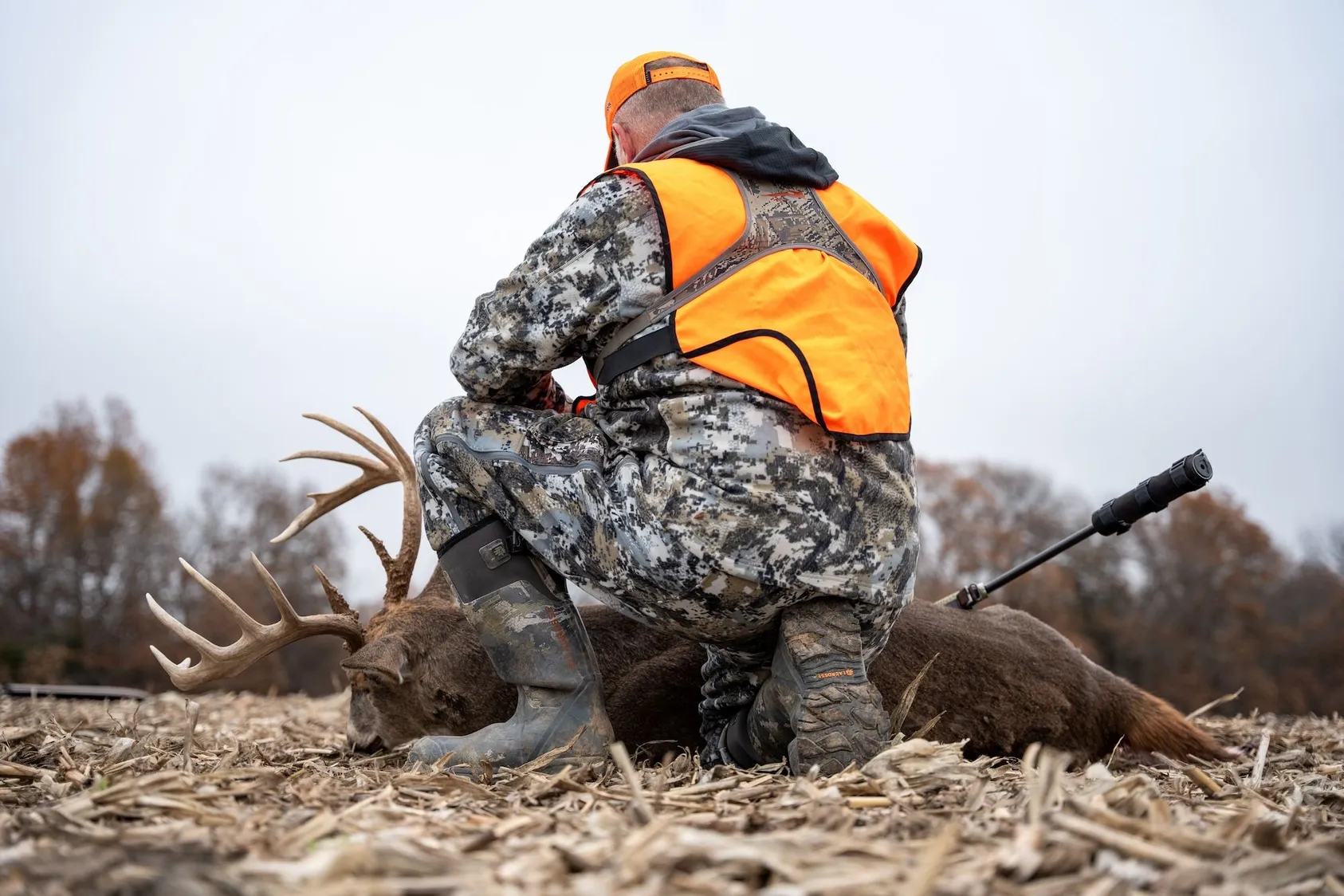 Hunter in orange safety vest and camouflage kneeling beside harvested deer in field, showing recreational hunting value of rural properties