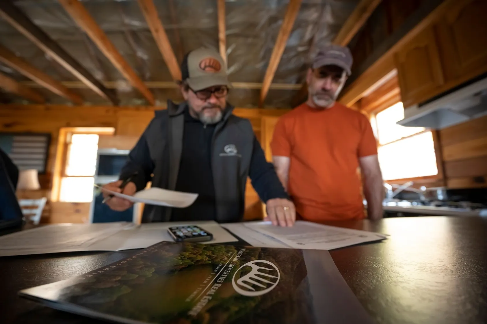 Two men in casual clothing reviewing documents at wooden table indoors, representing land buyers or investors discussing property financing options