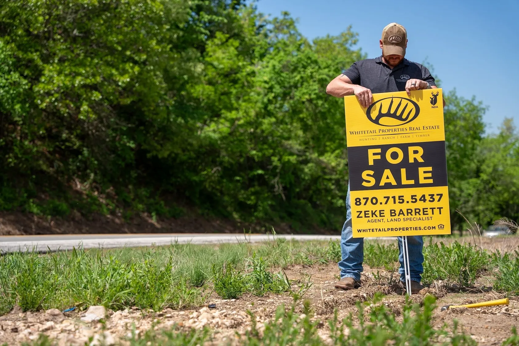 Real estate professional holding yellow 'For Sale' sign in rural outdoor setting, advertising available land for purchase by potential buyers