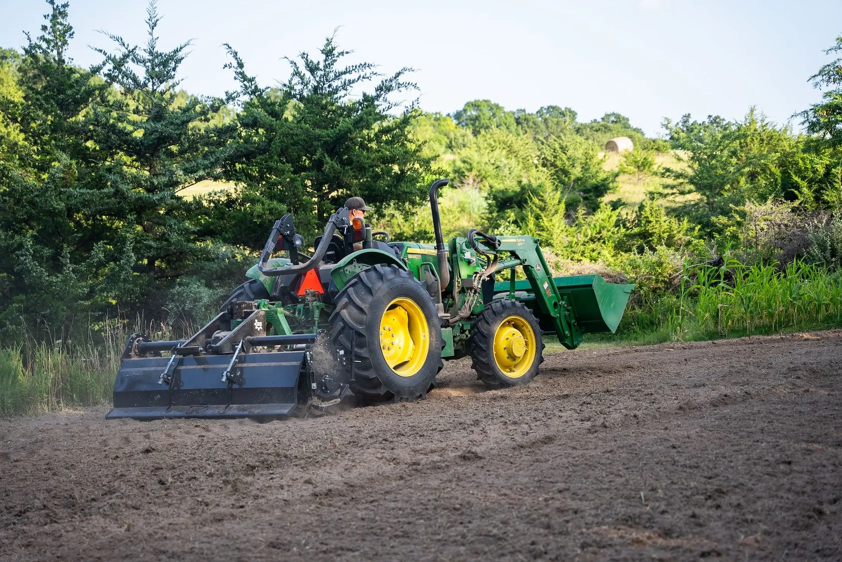 Green John Deere tractor with front loader and brush hog attachment working in rural field, demonstrating land clearing and maintenance equipment used in property development