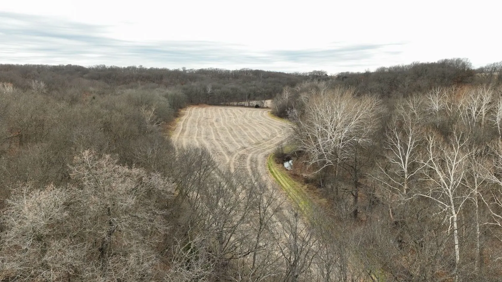 Aerial view of agricultural field with tractor creating dust cloud during farming operations, surrounded by wooded areas