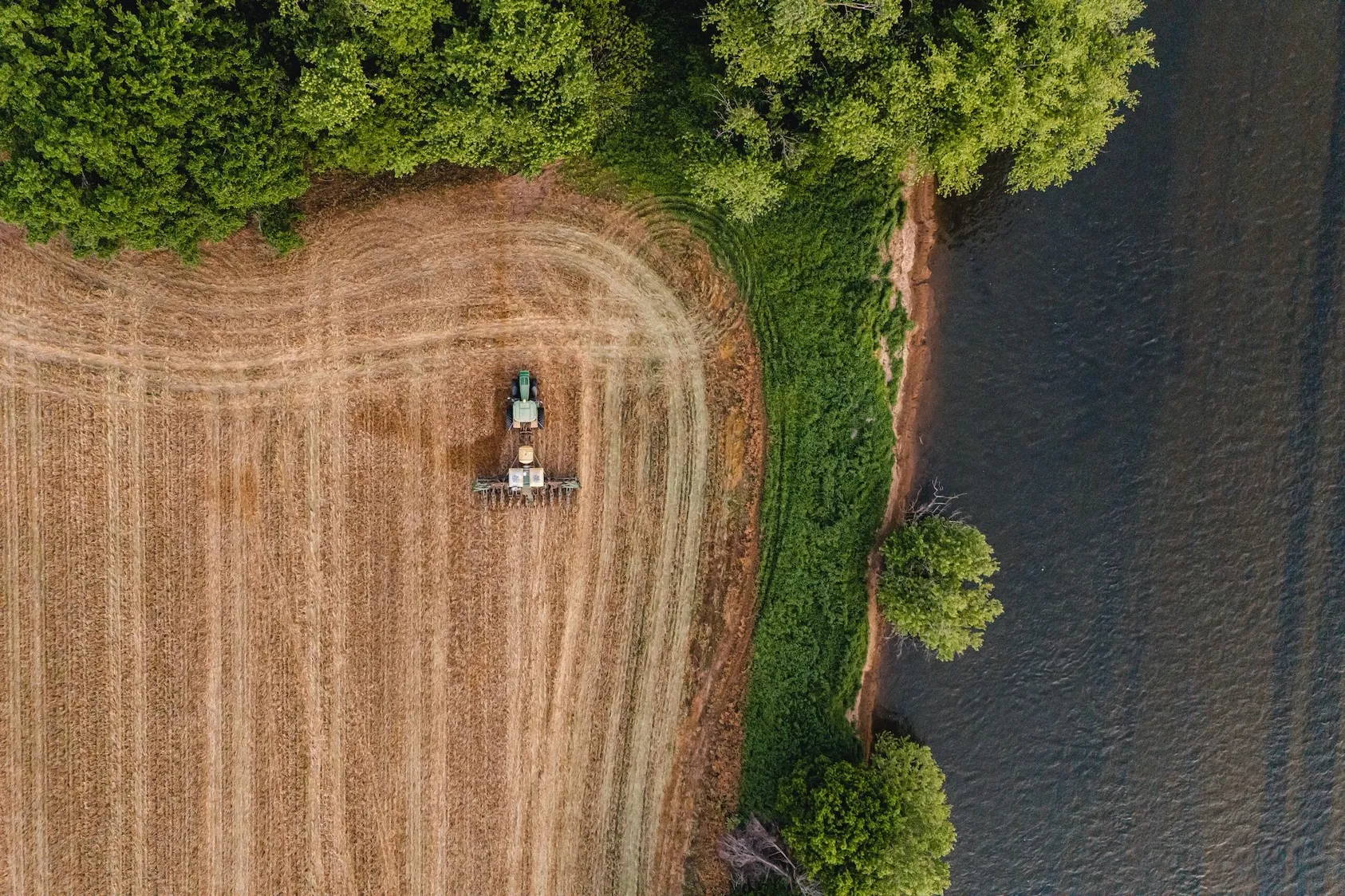 Aerial view of farming equipment working in golden agricultural field adjacent to wooded area, illustrating productive farmland and mixed-use property potential