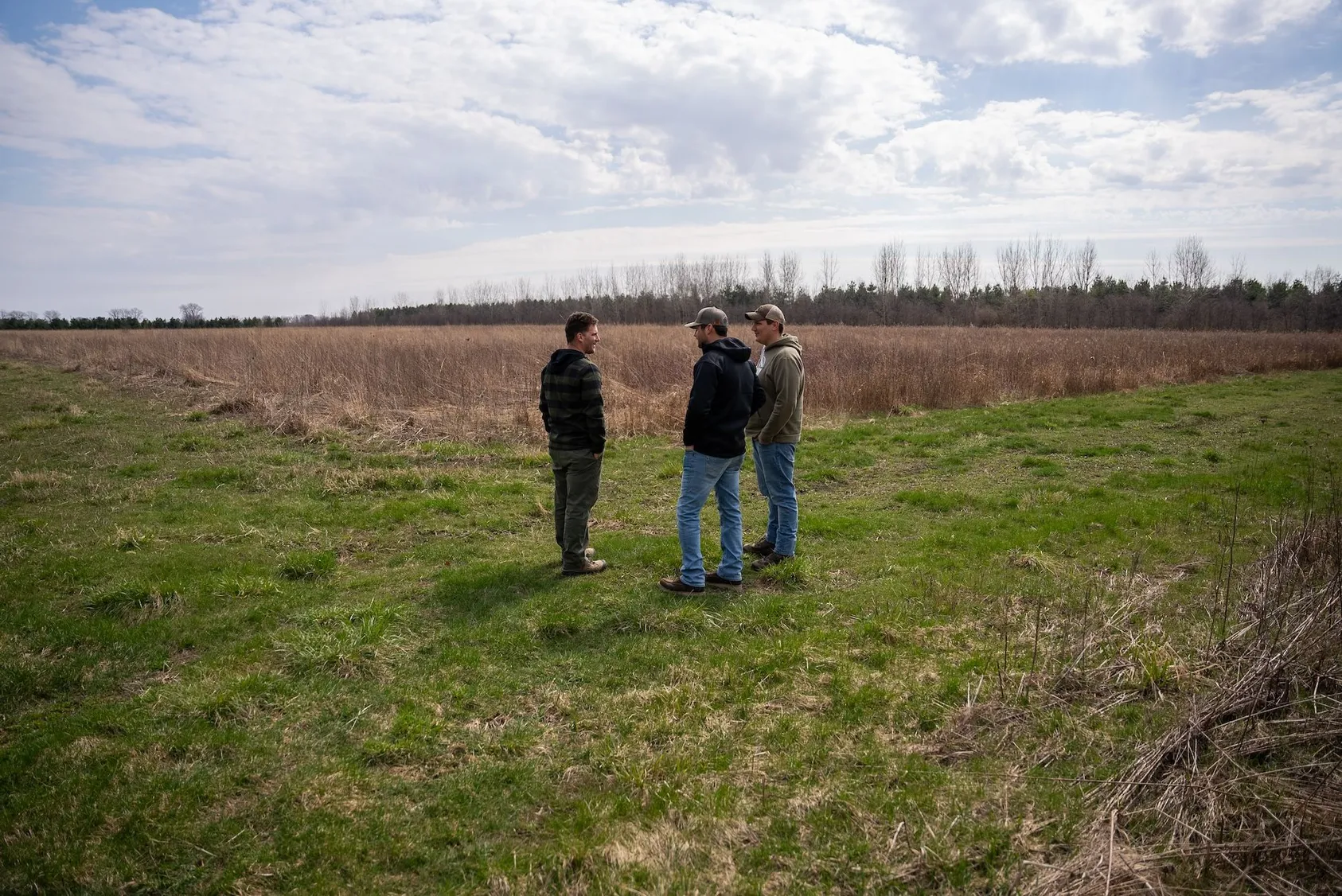 Three men standing and discussing in open rural field under cloudy sky, demonstrating due diligence process and property evaluation by potential buyers