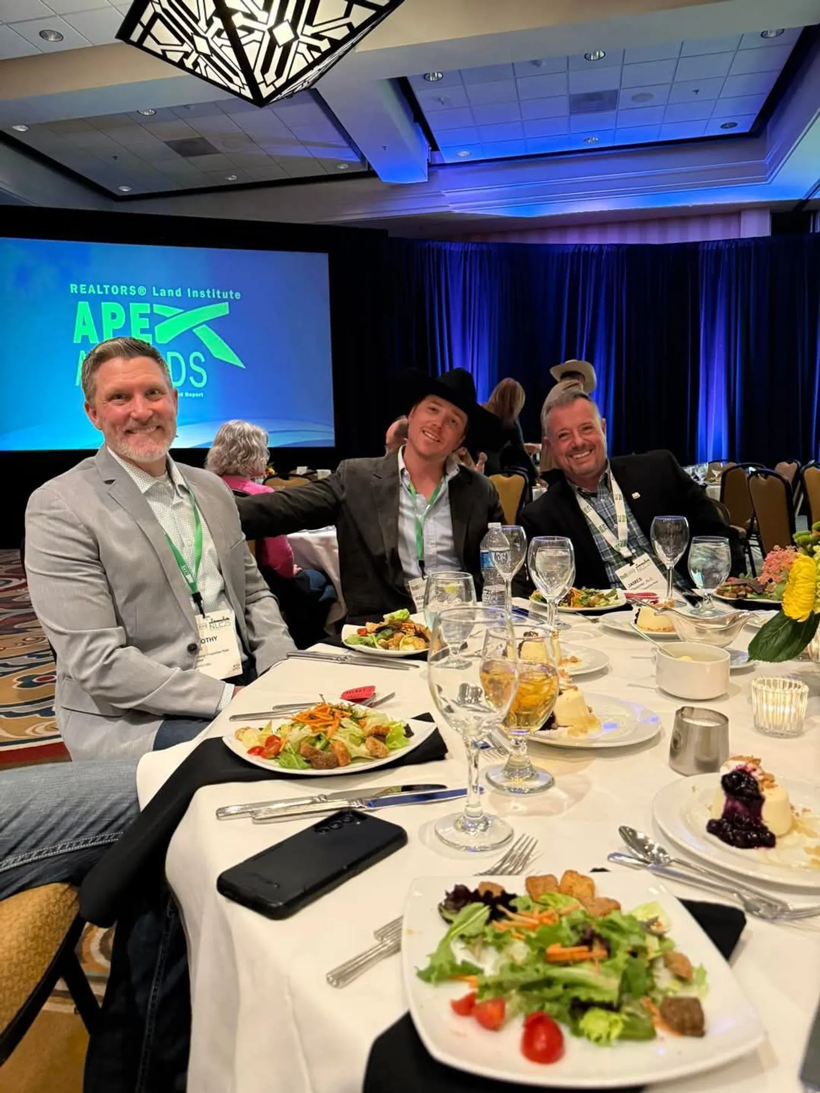 Three men in business suits sitting at a round dinner table during the RLI APEX Awards ceremony, with presentation screen visible in background