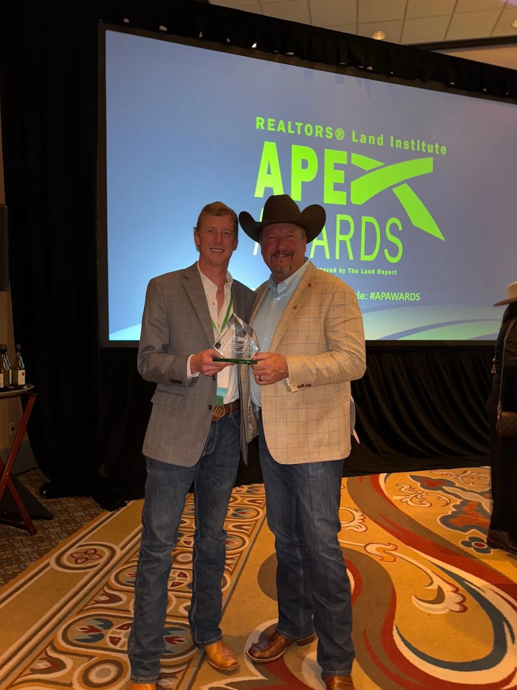 Two men in business suits on stage at APEX Awards ceremony, one presenting an award to the other with APEX Awards logo displayed on screen behind them