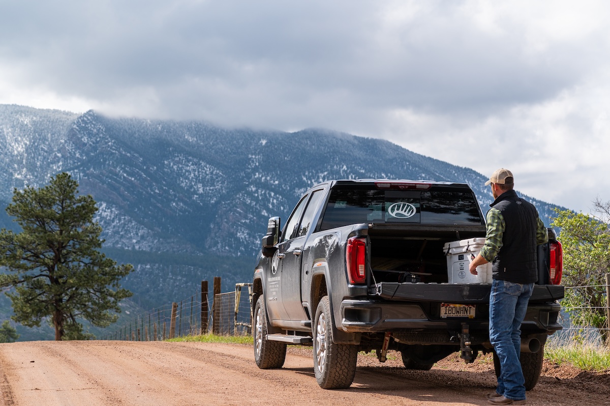 A man stands next to an open pickup truck on a dirt road, with snow-capped mountains and a tree in the background under a cloudy sky.
